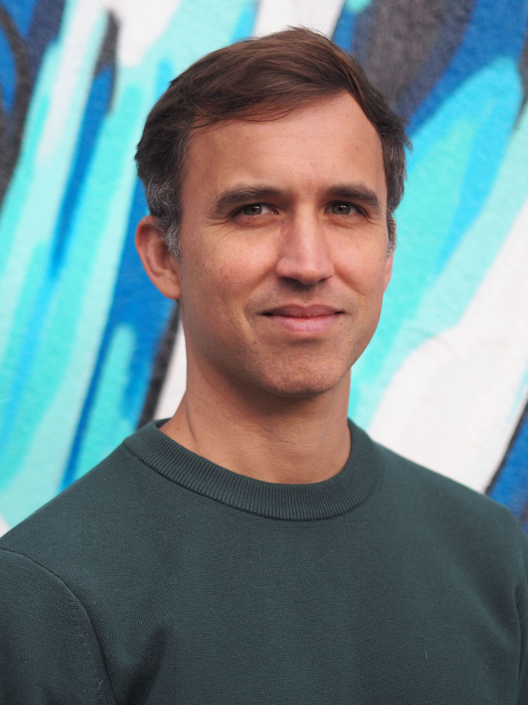 Portrait of Andre DePonti in front of a blue mural background, wearing a dark green sweater and looking at the camera.
