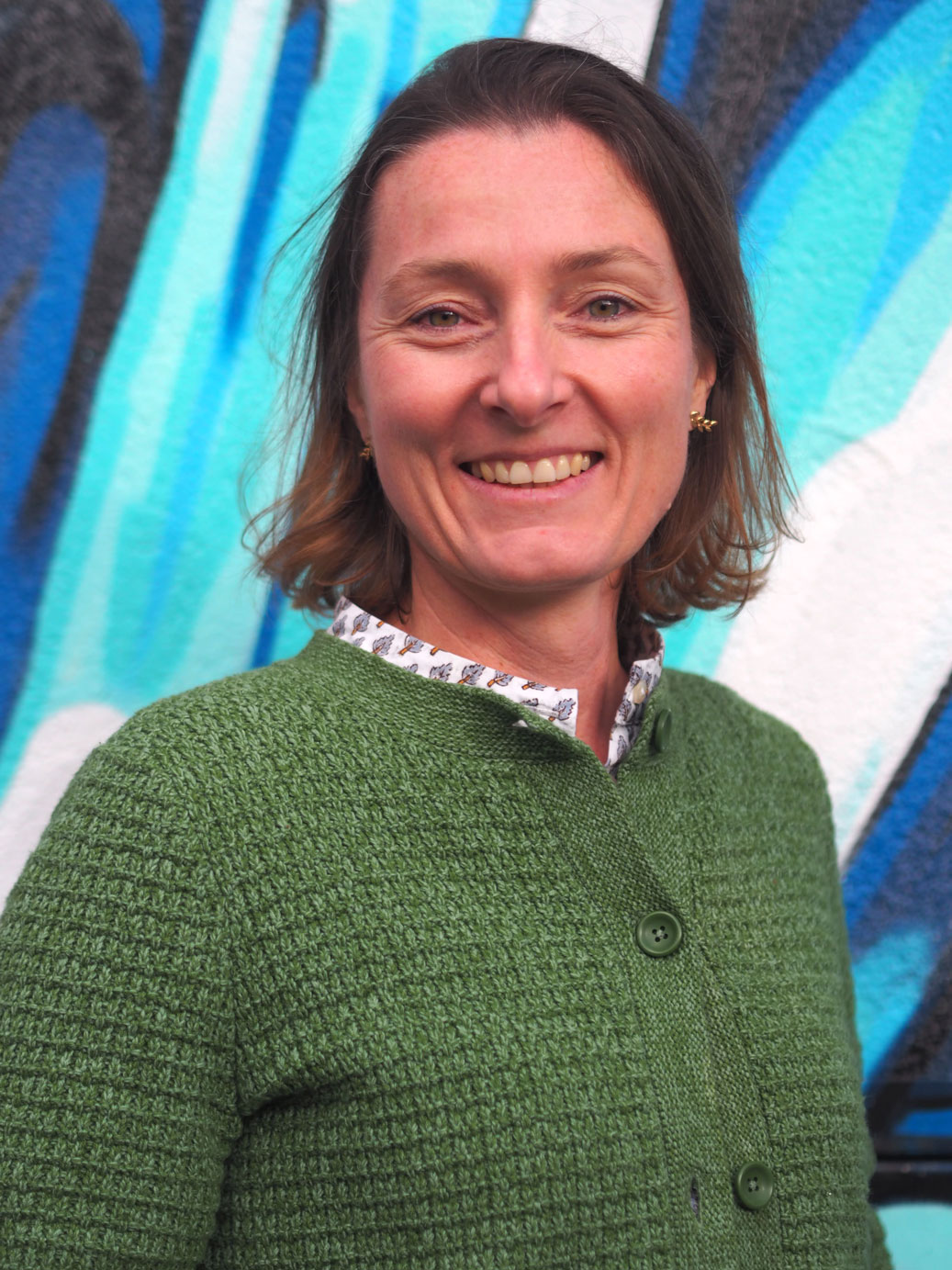 Portrait of Maite Matos Ichaso smiling in front of a blue mural background, wearing a green knitted cardigan.