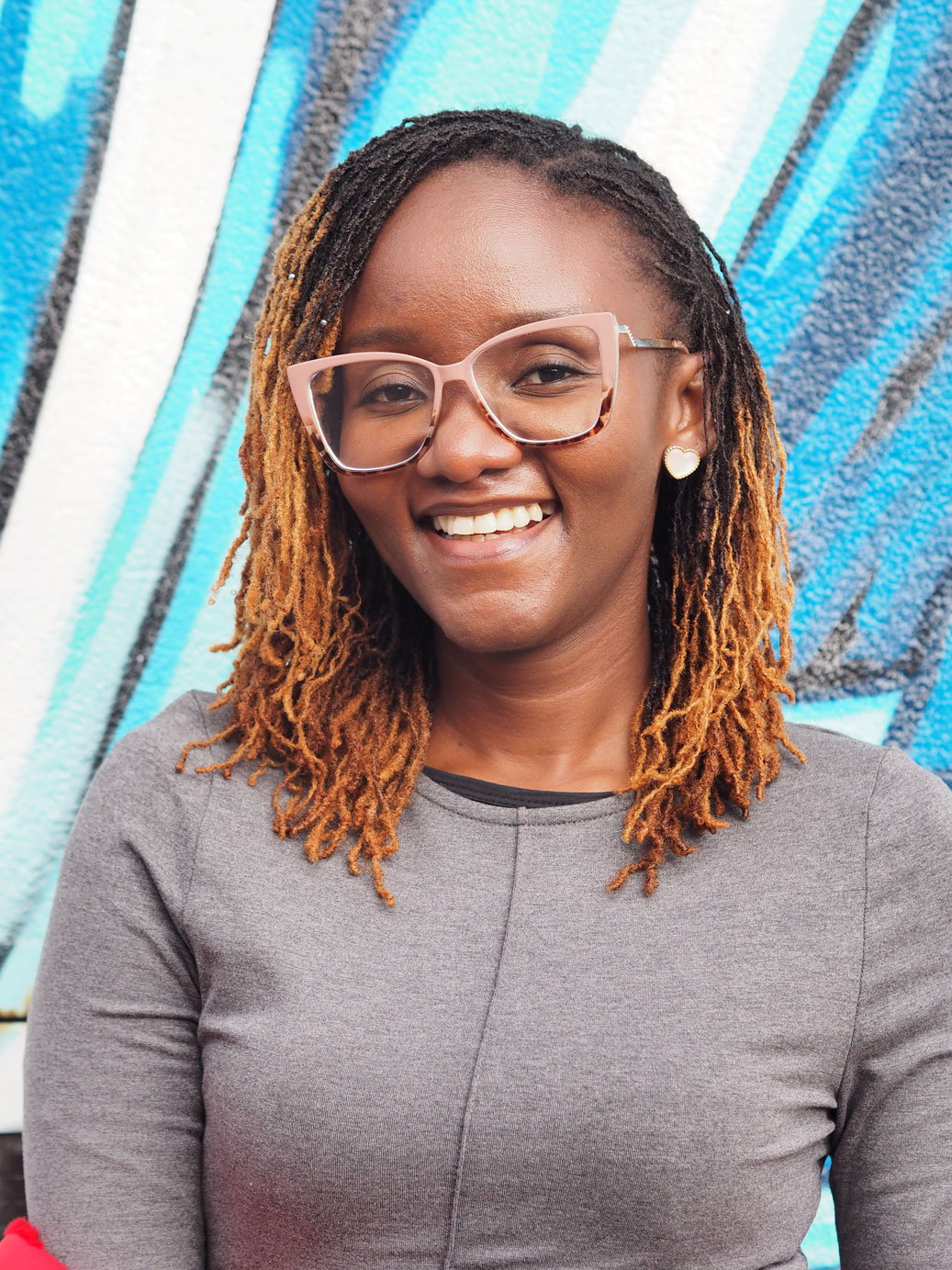Portrait of Phonsina Archane smiling in front of a blue mural background, wearing pink glasses and a grey top.
